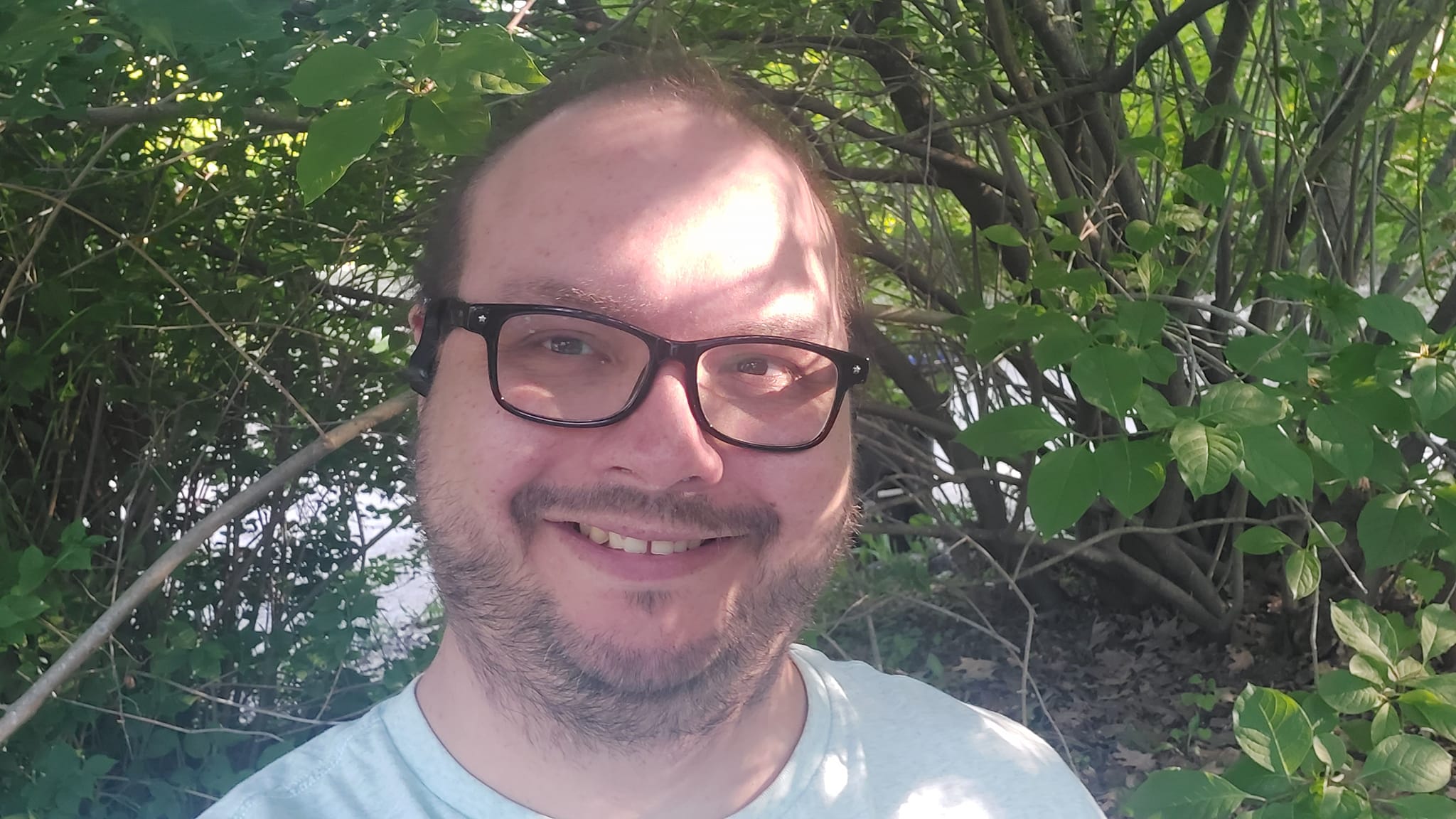 A man wearing a blue shirt stands in front of a bush smiling